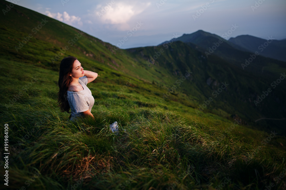 A girl sits on the edge of the cliff and looking at the sun valley and mountains. Woman sitting on mountain top and contemplating the sunset. Young woman in long white dress