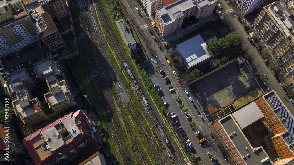 Aerial perpendicular view of the tracks of a train station passing by a road where there are cars parked. A few bushes of grass grows between the rails. There is nobody.