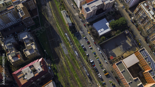 Aerial perpendicular view of the tracks of a train station passing by a road where there are cars parked. A few bushes of grass grows between the rails. There is nobody.
