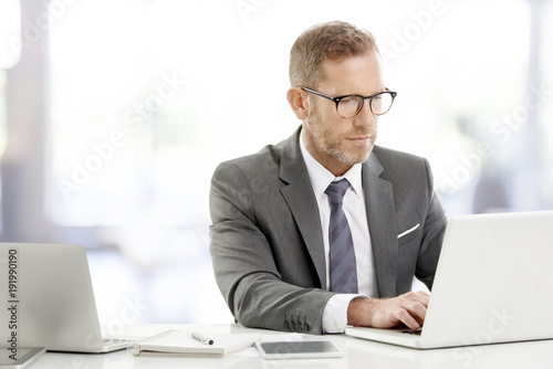 Middle aged businessman with laptop. Portrait of mature senior male agent wearing shirt and tie while sitting at desk and working on laptop at the office.  © gzorgz