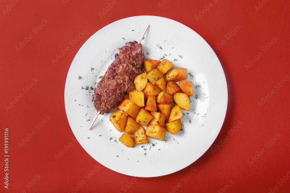 Meat kebab from pork, beef with fried potatoes on a white plate. Red monophonic background. A hearty meal, dinner. Dish for the menu. View from above Meat kebab from pork, beef with fried potatoes on a white plate. Red monophonic background. A hearty meal, dinner. Dish for the menu. View from above