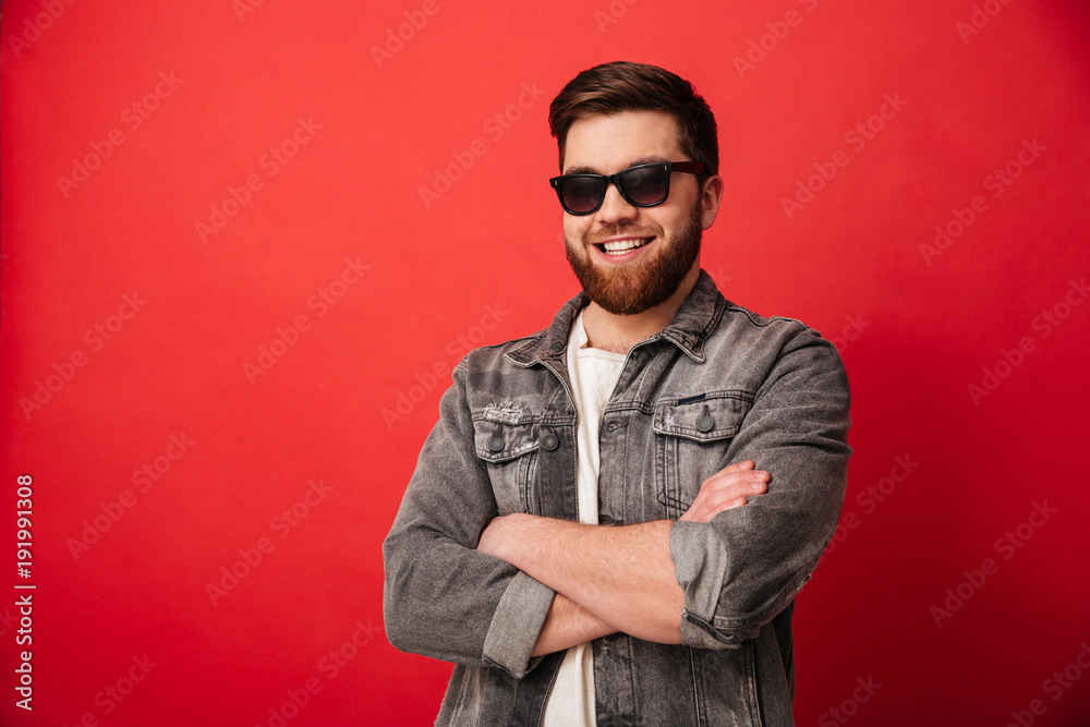 Plakat Portrait of attractive man 30s in sunglasses smiling on camera and posing with hands folded, isolated over red background