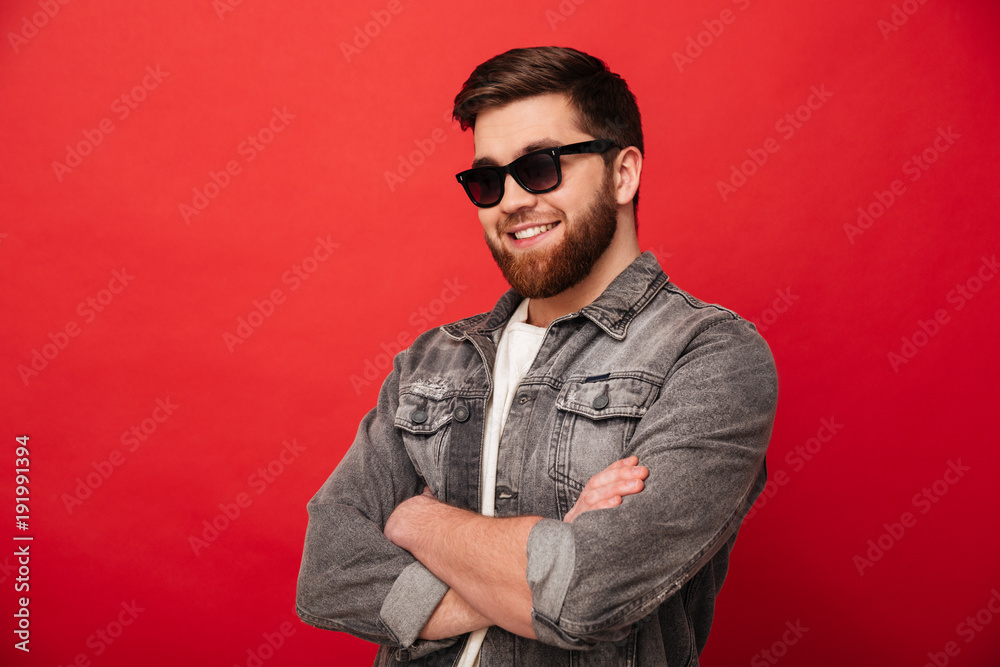 Plakat Portrait of irresistible stylish man 30s in sunglasses smiling on camera and posing with hands crossed, isolated over red background