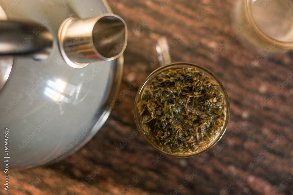 Making tea (green tea leaves) in a glass cup, wit two pieces, green tea leaves with hot water in a transparent clear cup with a kettle near it, making tea with kettle Making tea (green tea leaves) in a glass cup, wit two pieces, green tea leaves with hot water in a transparent clear cup with a kettle near it, making tea with kettle