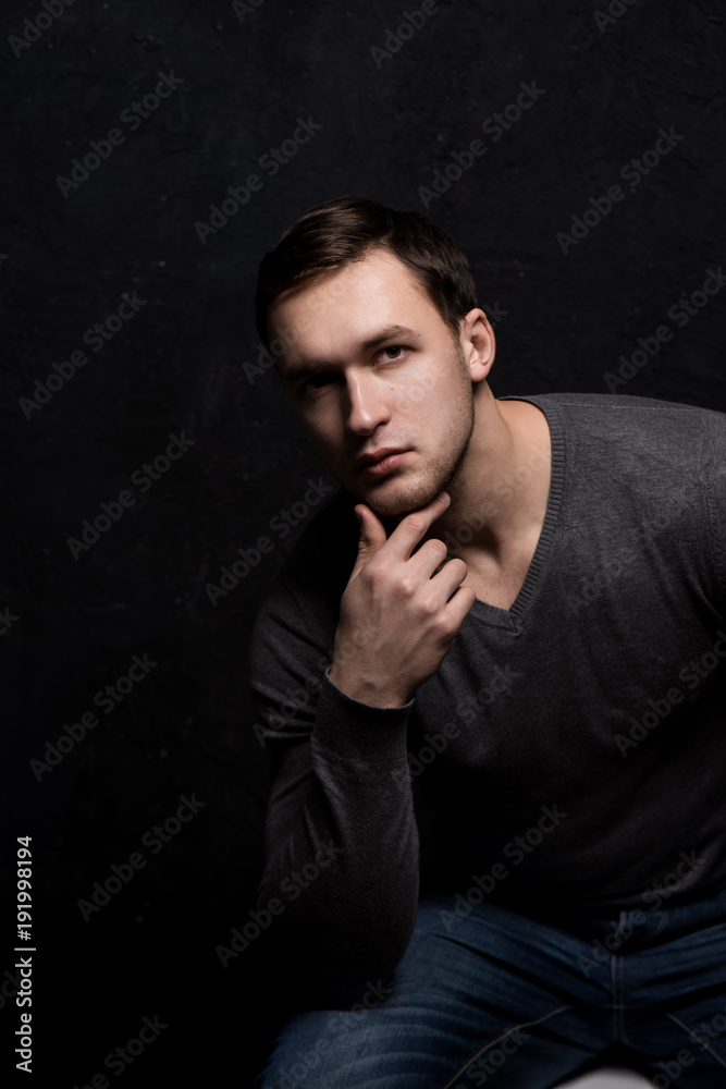 Handsome tough guy on a black background. Hand at the face. Thoughtfulness. Portrait. Brawn. Gray sweater. Force. Masculinity.