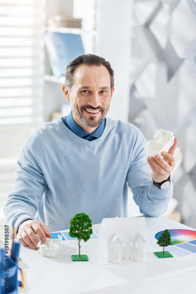 Being an architect. Good-looking cheerful dark-haired bearded man smiling and holding a model of a future house and sitting at the table with other models Being an architect. Good-looking cheerful dark-haired bearded man smiling and holding a model of a future house and sitting at the table with other models