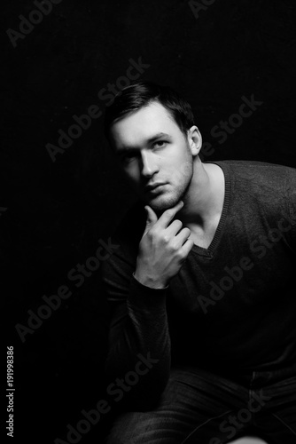 Handsome tough guy on a black background. Hand at the face. Black and white photo. Thoughtfulness. Portrait. Brawn. Gray sweater. Force. Masculinity.