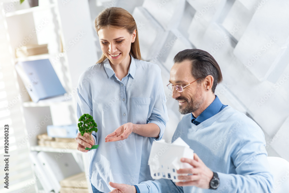 Obraz premium Nice atmosphere. Attractive happy fair-haired young woman smiling and working with her colleague holding a house miniature and holding a tree miniature