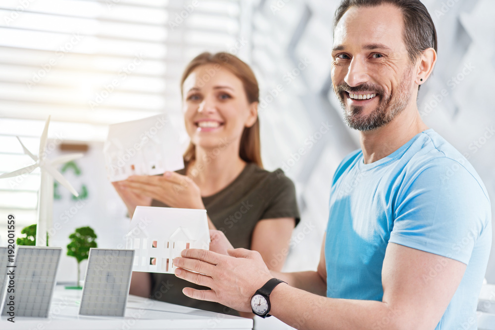 Great team. Handsome glad dark-haired bearded man smiling and holding a house miniature and his colleague sitting in the background smiling Great team. Handsome glad dark-haired bearded man smiling and holding a house miniature and his colleague sitting in the background smiling