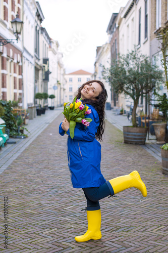 girl with a bouquet of spring tulips in pink. In a blue raincoat and yellow boots on the street in Europe. Happy woman. Smile on the face. Card.