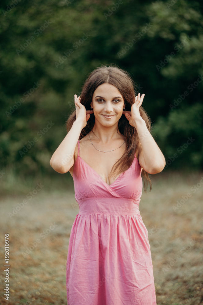 Beautiful slender brunette woman in short gently pink dress is standing in a forest. The woman straightens her hair. She puts her hair in pigtails. Beautiful slender brunette woman in short gently pink dress is standing in a forest. The woman straightens her hair. She puts her hair in pigtails.