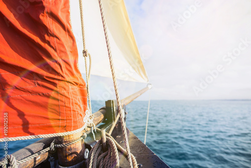 rope wound on a wooden cleat fixed on the hull of a rigging vintage sailing boat with a beige jib and an ocher sail filled by the wind at the blur background during a sunny sea trip in brittany