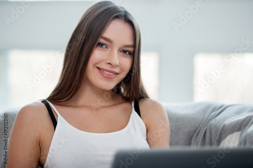 Inspired. Beautiful cheerful long-haired slim girl smiling and sitting on the sofa and wearing a top and relaxing