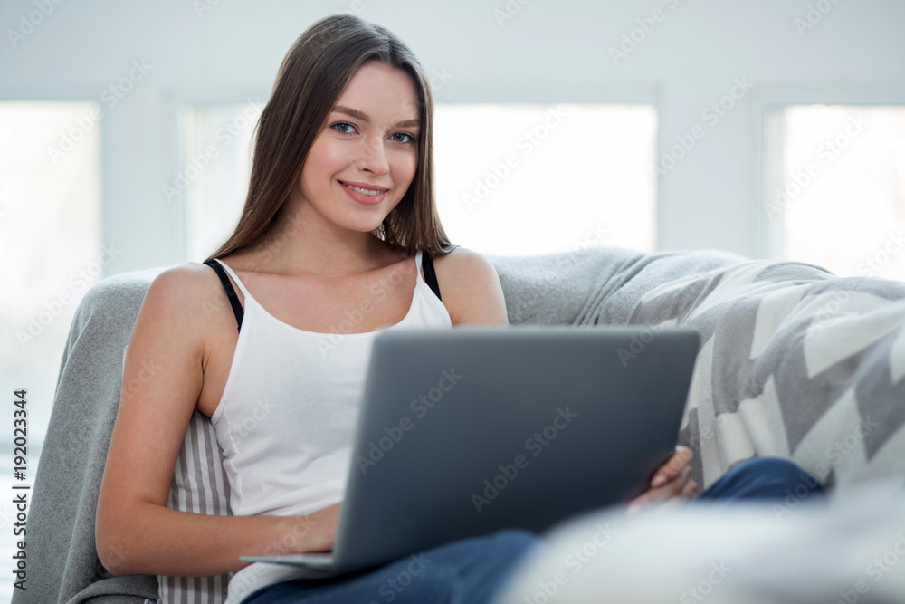 Distance working. Beautiful exuberant dark-haired young woman smiling and sitting on the sofa and working on her laptop Distance working. Beautiful exuberant dark-haired young woman smiling and sitting on the sofa and working on her laptop