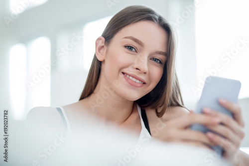 Being delighted. Good-looking exuberant dark-haired young woman smiling and wearing a top and holding her phone