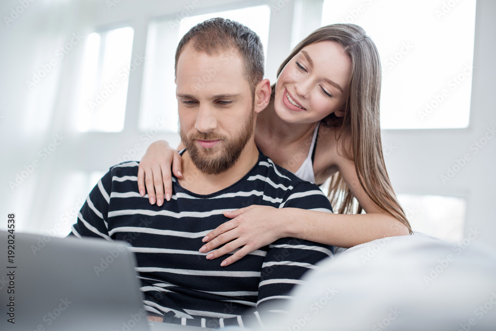 I treasure you. Pretty content long-haired young woman hugging her boyfriend and smiling while he using a laptop