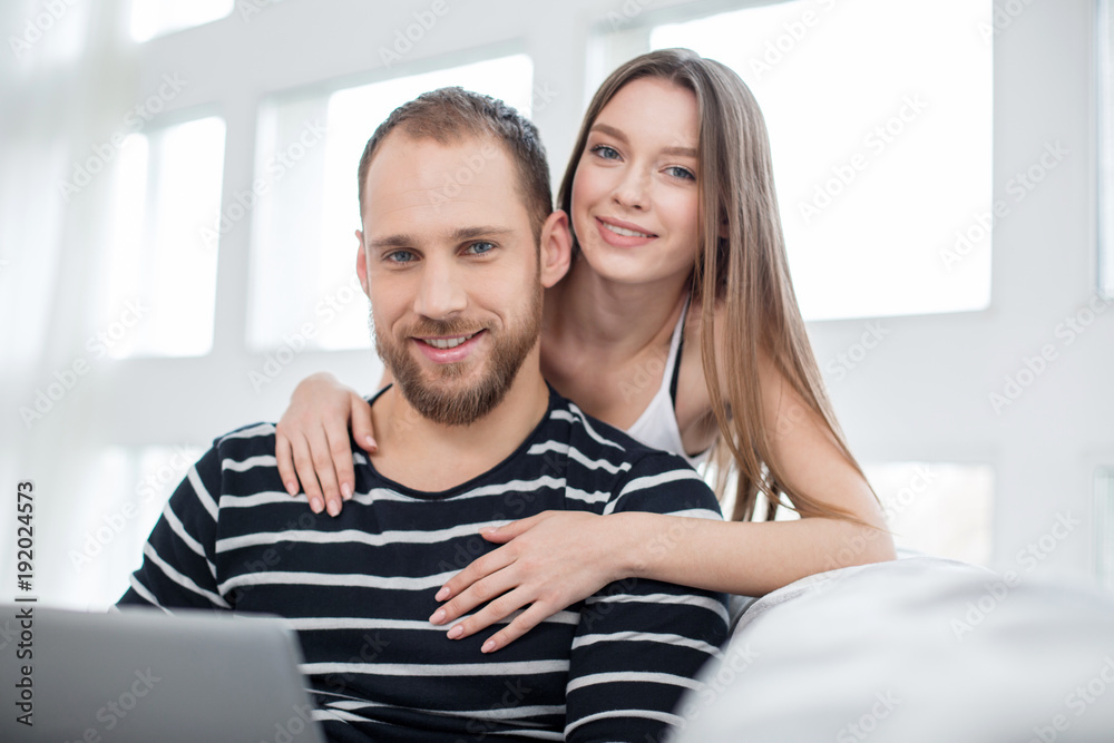 My beloved. Attractive glad long-haired young woman hugging her handsome boyfriend and smiling while he using a laptop My beloved. Attractive glad long-haired young woman hugging her handsome boyfriend and smiling while he using a laptop