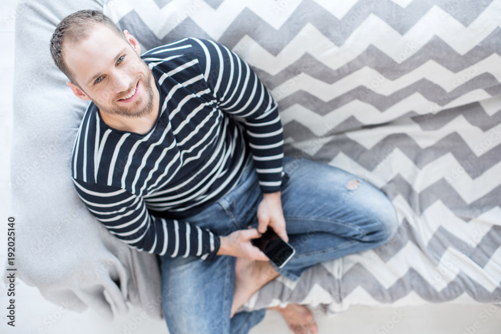 Being energetic. Good-looking cheerful bearded young man smiling and sitting on the couch and holding his phone