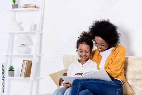 happy african american mother and daughter reading book together at home