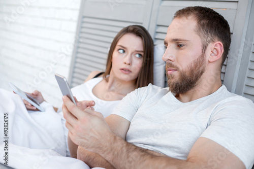 Checking his messages. Pretty unsmiling dark-haired young woman holding her phone and looking at her man typing on his phone while he lying next to her