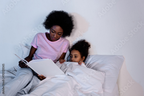 african american mother reading book for daughter in evening at home