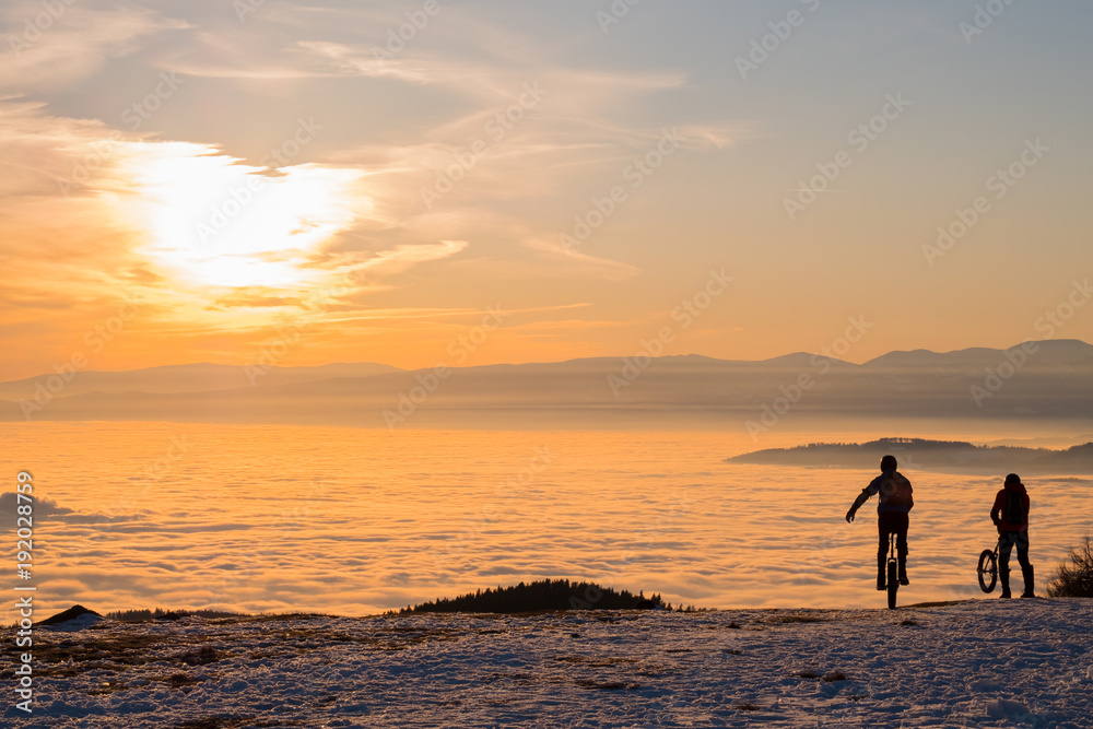 Men on unicycles riding on mountain over fog to sunset