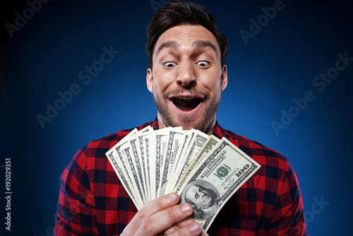 Handsome bearded man in red shirt. Funny guy is a lucky winner, she is holding a pile of money, he is surprised and can't believe it, he is happy to win one million dollar jackpot, now he is rich.