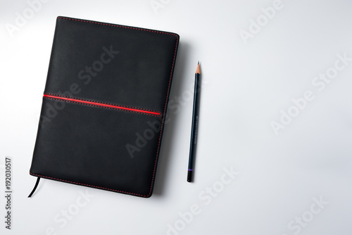 Top view blank black leather diary and pencil on white desk.