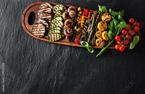 a large portion of colored grilled vegetables and mushrooms on a wooden tray on a dark slate. festive summer food