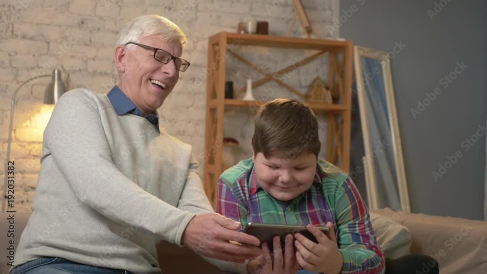 Grandfather and grandson sitting on the couch using a tablet watching a funny movie, laughing, smiling. Home comfort, family idyll, cosiness concept 60 fps Grandfather and grandson sitting on the couch using a tablet watching a funny movie, laughing, smiling. Home comfort, family idyll, cosiness concept 60 fps