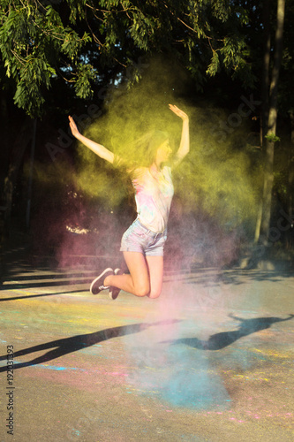 Splendid brunette woman jumping with exploding pink  blue and yellow Holi powder in the park
