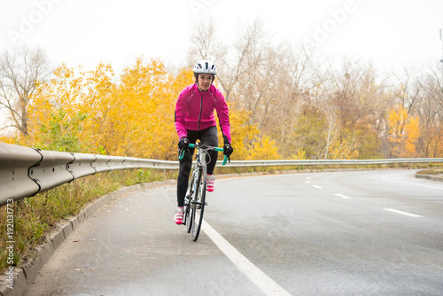 Fototapeta Naklejka Na Ścianę i Meble -  Young Woman in Pink Jacket Riding Road Bicycle on the Highway in the Cold Autumn Day. Healthy Lifestyle.