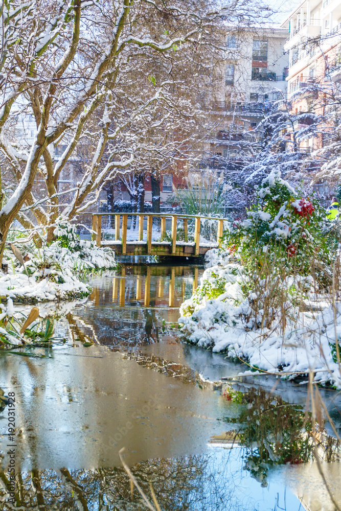 Small river with mini pont in the park, outdoor in Paris, France, during snowfall in winter. Taken in the morning with sunrise light. Smart concept of modern park for family recreation.