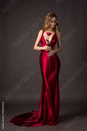 Beautiful blonde girl wearing a deep neckline red satin dress holding red rose in her hand on a grey background