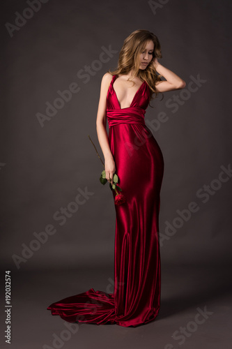 Beautiful blonde girl wearing a deep neckline red satin dress holding red rose in her hand on a grey background