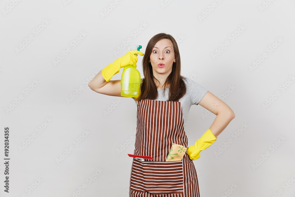 Young fun housewife in yellow gloves, striped apron, cleaning rag, squeegee in pocket isolated on white background. Housekeeper woman shoots from spray bottle with cleaner liquid. Bottle copy space. Young fun housewife in yellow gloves, striped apron, cleaning rag, squeegee in pocket isolated on white background. Housekeeper woman shoots from spray bottle with cleaner liquid. Bottle copy space.