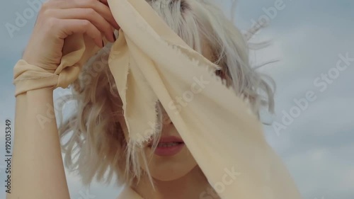 Portrait of a charming young woman posing on a cloudy day on nature. She is hiding herself behind silk cloth.