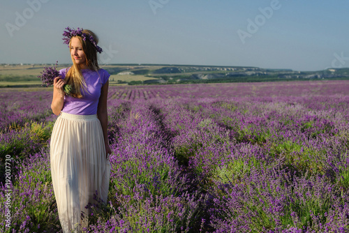 Charming girl in a hat and a light dress posing in a lavender field at sunset