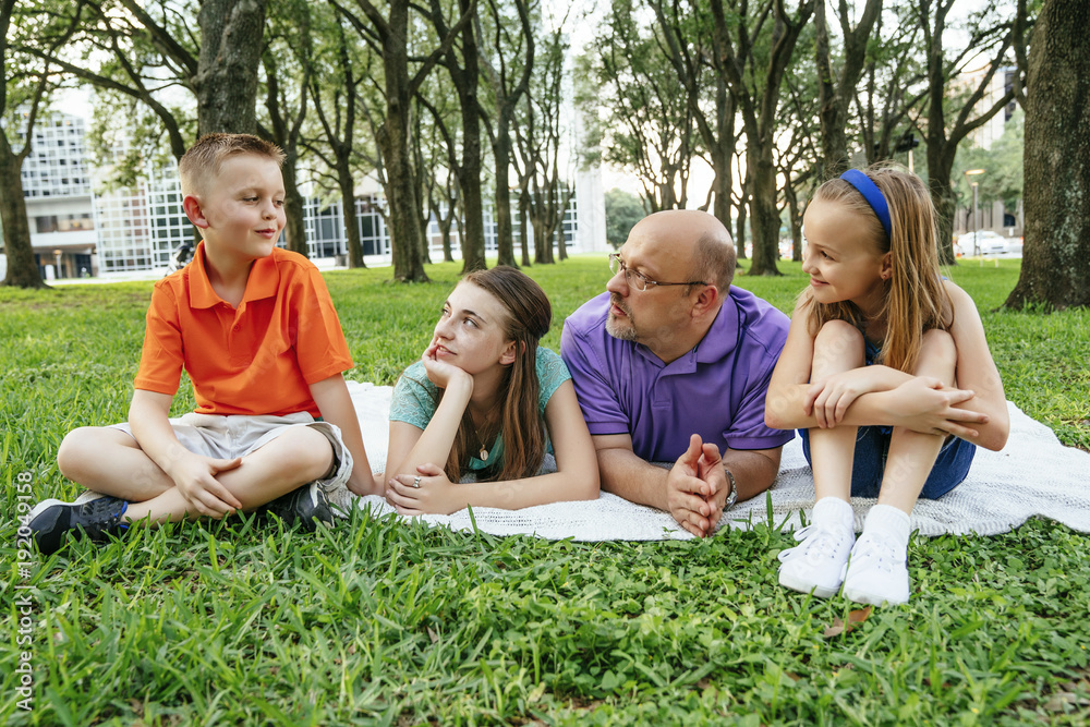 Caucasian father and daughters laying on blanket in park listening to boy Caucasian father and daughters laying on blanket in park listening to boy