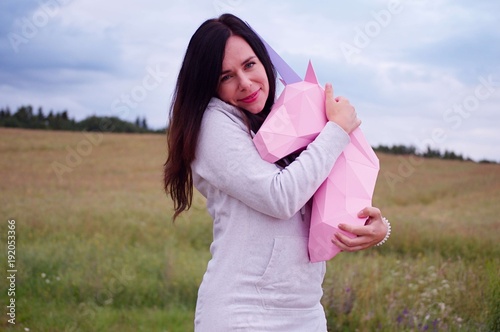 Beautiful happy smiling young girl with pink paper unicorn. Meadow and blue sky on the backgroundSummer, field, nature photo