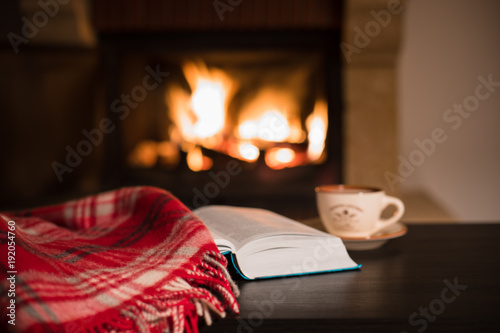 Wool warm blanket or plaid and a book with a cup of tea on the background of burning fireplace