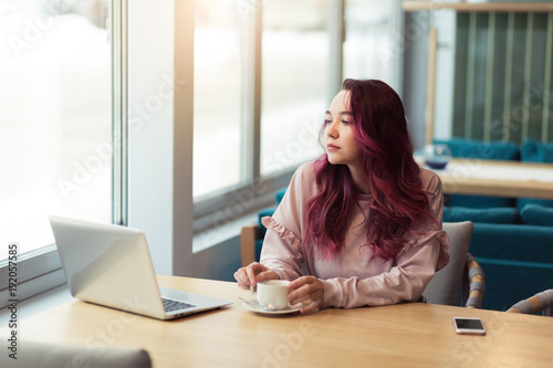 Young hipster girl works at laptop in a cafe, typing on the keyboard. Workplace in a modern sunny cafe, a cup of tea near a laptop