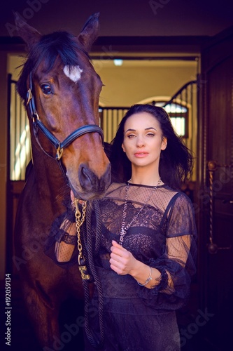 Gorgeous stylishly dressed woman in dark lace boho style dress and gold jewelry with a thoroughbred horse. The concept of love human and animals, horses and rider lifestyle 