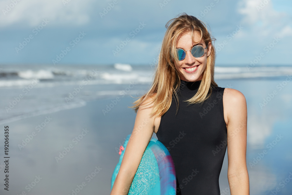Watersport summer activities and recreation concept. Smiling beautiful young woman in shades and swimsuit, holds surfboard, poses against ocean background, being in good mood as likes her hobby Watersport summer activities and recreation concept. Smiling beautiful young woman in shades and swimsuit, holds surfboard, poses against ocean background, being in good mood as likes her hobby