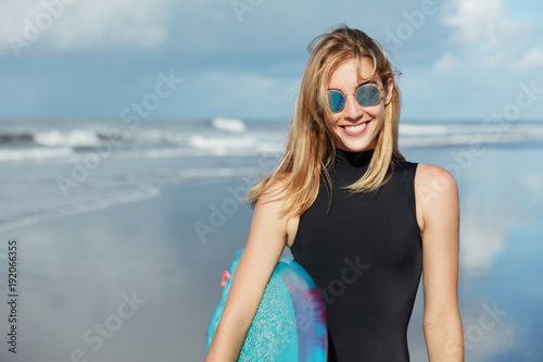 Watersport summer activities and recreation concept. Smiling beautiful young woman in shades and swimsuit, holds surfboard, poses against ocean background, being in good mood as likes her hobby