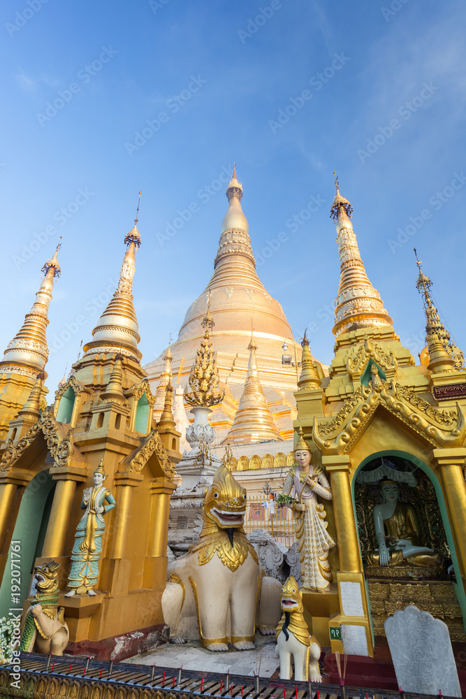 Small pagodas and statues in front of the gilded Shwedagon Pagoda in Yangon, Myanmar on a sunny day.
