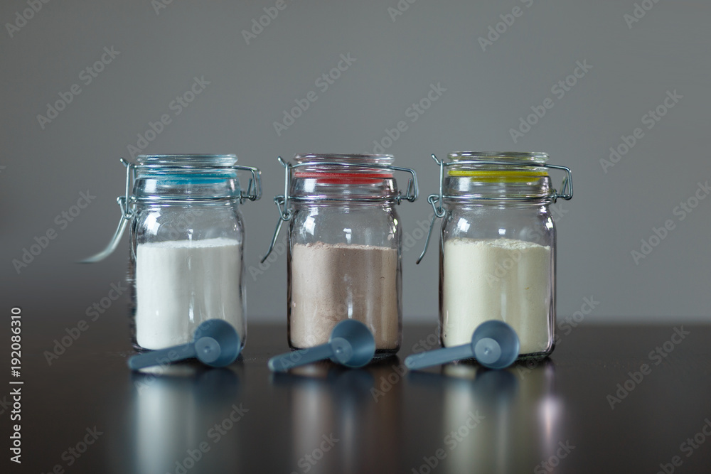 Three glass jars with protein powder, strawberries, banana and chocolate and three scoops for milkshakes. Sports and dietary nutrition for athletes and weight loss or muscle mass bodybuilders. Three glass jars with protein powder, strawberries, banana and chocolate and three scoops for milkshakes. Sports and dietary nutrition for athletes and weight loss or muscle mass bodybuilders.