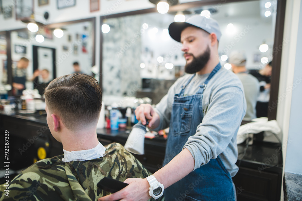 A stylish barber with a beard finishes a male hairstyle to the client. Men's haircuts in barbershop. Young handsome barber making haircut of attractive man in barbershop A stylish barber with a beard finishes a male hairstyle to the client. Men's haircuts in barbershop. Young handsome barber making haircut of attractive man in barbershop