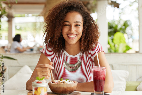 Adorabe dark skinned female with Afro hairstyle, wears casual striped t shirt, eats exotic sweet dish, drinks cocktail at outdoor cafeteria, smiles broadly, demonstrates white teeth, rests after work photo