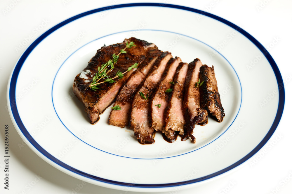 10oz Ribeye steak with thyme and smokey applewood seasoning Sliced and fanned on a blue rim plate on a white background With fork and steak knife